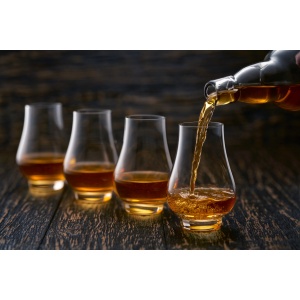 Whisky being poured from a bottle into a row of four nosing glasses on a dark wooden table