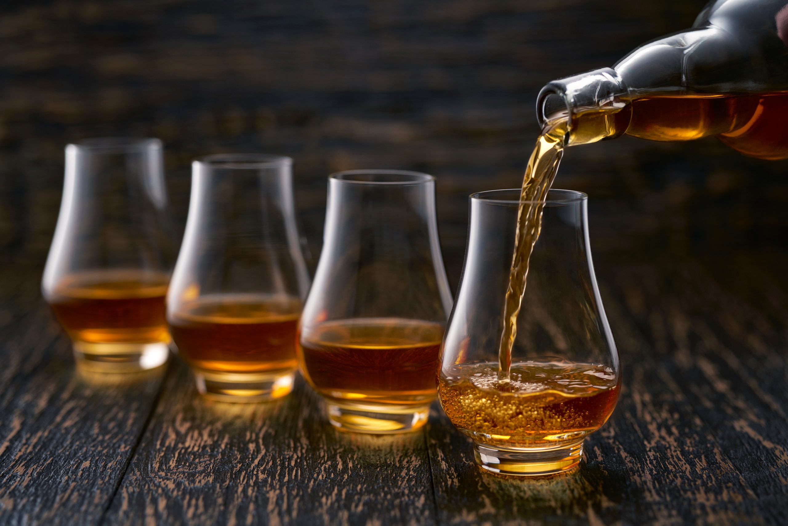 Whisky being poured from a bottle into a row of four nosing glasses on a dark wooden table